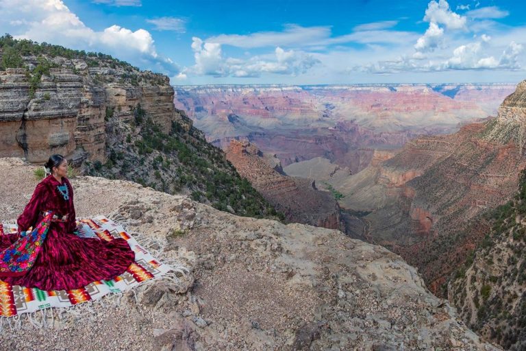 A Diné woman in a traditional dress, on the rim of Grand Canyon. Image courtesy of Grand Canyon National Park via via Flickr (CC BY-NC-ND 2.0).