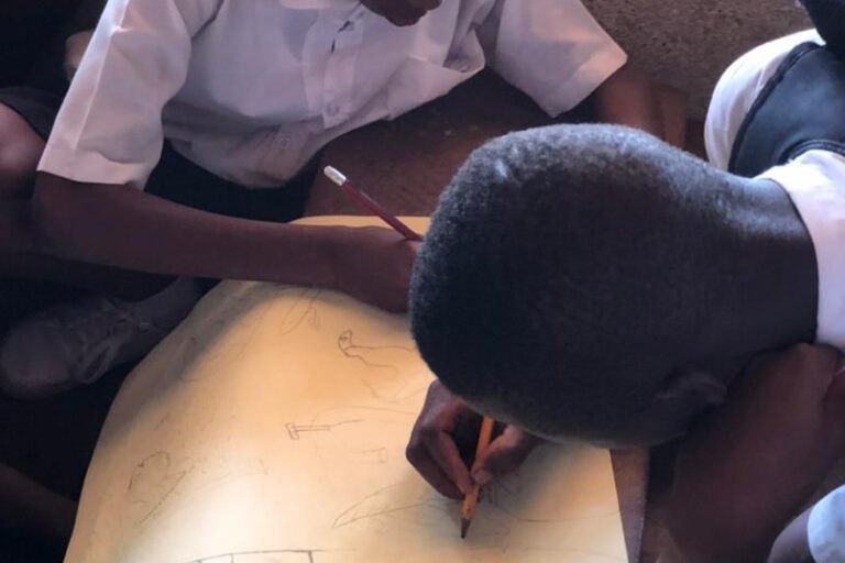 Two students bent over a drawing of a house and villagers. Image courtesy Sierra Leone Environment Matters.