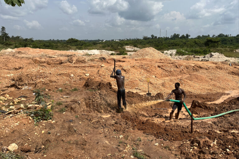 A man standing amidst mining pits raises a pickaxe; his companion directs dirty water from a hose towards his feet. Image by Élodie Toto for Mongabay.