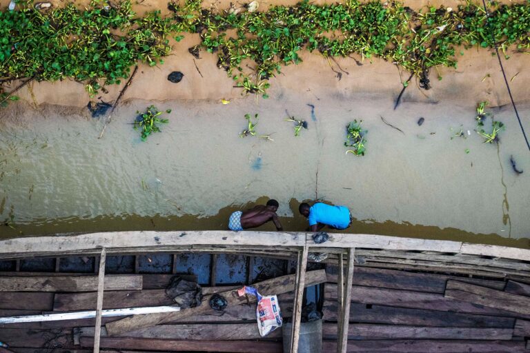 Overhead shot of two men standing in shallow water, bent over repairing a large wooden canoe. Image by Bolaji Eiseke via Wikicommons (CC BY-SA 4.0)