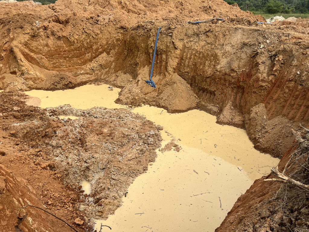 Muddy yellow water against the red earth of a mining pit near Kumasi, Ghana, an abandoned shovel just upright from soil on the far side. Image by Élodie Toto for Mongabay.