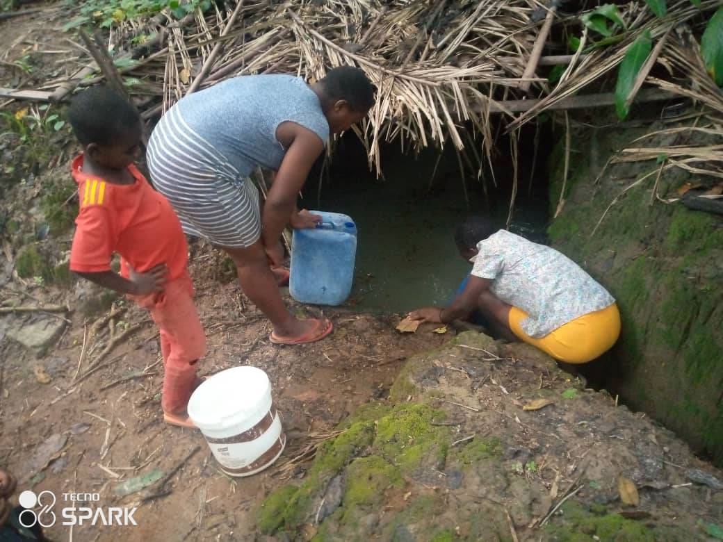 A woman with two younger children filling plastic containers from a murky pool of water, partly shaded by a rough screen of palm fronds. Image courtesty Suhucam.