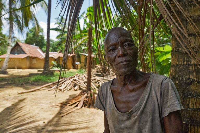 A thin elderly man, the open neck of hard-worn brown t-shirt exposing bones in his chest, looks at the camera, the trunk of a palm tree, dried fronds, and simple houses with rusting corrugated iron roofs in the background. Image by Ashoka Mukpo for Mongabay.